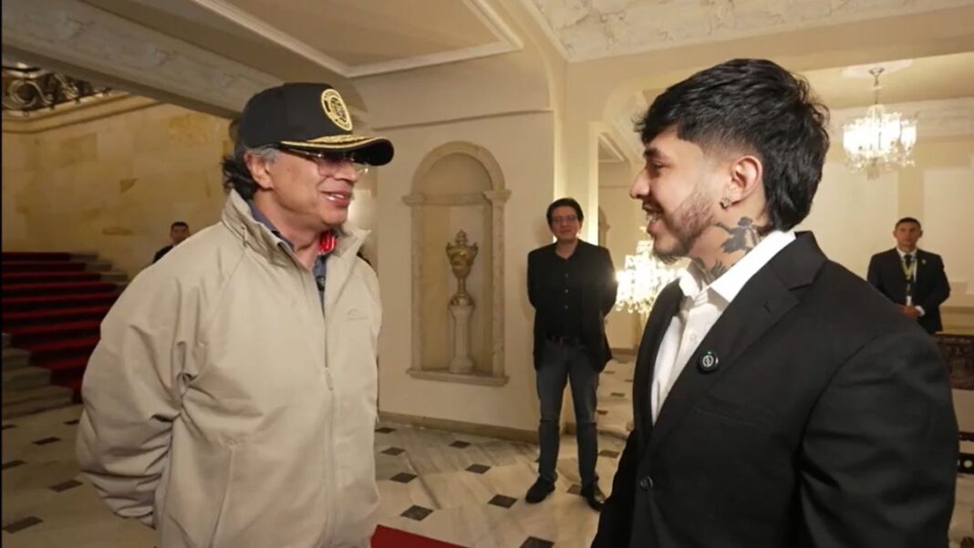 Fotografía del presidente de Colombia, Gustavo Petro, usando gorra negra y chaqueta clara, sonriendo de frente al popular streamer Westcol, quien viste un traje negro formal que deja ver los tatuajes de su cuello. Ambos conversan amistosamente en los elegantes pasillos de la Casa de Nariño durante su polémica entrevista transmitida en vivo.