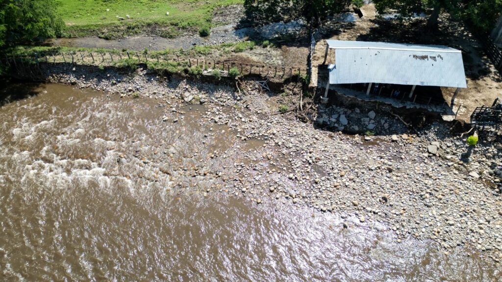 Vista aérea de la erosión en la ribera de un río y daños en una estructura con techo de zinc debido al aumento del caudal por lluvias.