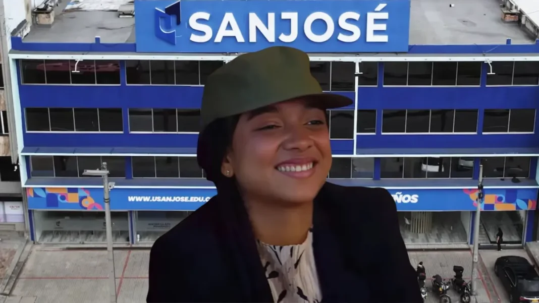 Una fotografía de Juliana Guerrero, exasesora imputada bajo cargos de fraude procesal relacionado con diplomas universitarios falsos (cartones chimbos), sonriendo ampliamente mientras usa una gorra militar verde y una chaqueta negra. La imagen la muestra frente al edificio de la Fundación Universitaria San José (SANJOSE), de donde presuntamente provienen los títulos falsificados que usó para postularse a viceministra de las juventudes.