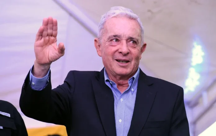 Fotografía de medio cuerpo del expresidente de Colombia, Álvaro Uribe Vélez, levantando su mano derecha en señal de saludo durante un evento público.