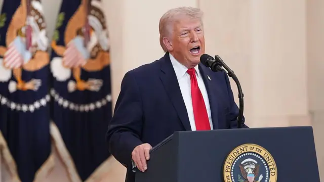 Primer plano de Donald Trump gritando con la boca abierta durante un discurso apasionado en un podio oficial que muestra el sello del Presidente de los Estados Unidos. Dos banderas estadounidenses en el fondo.