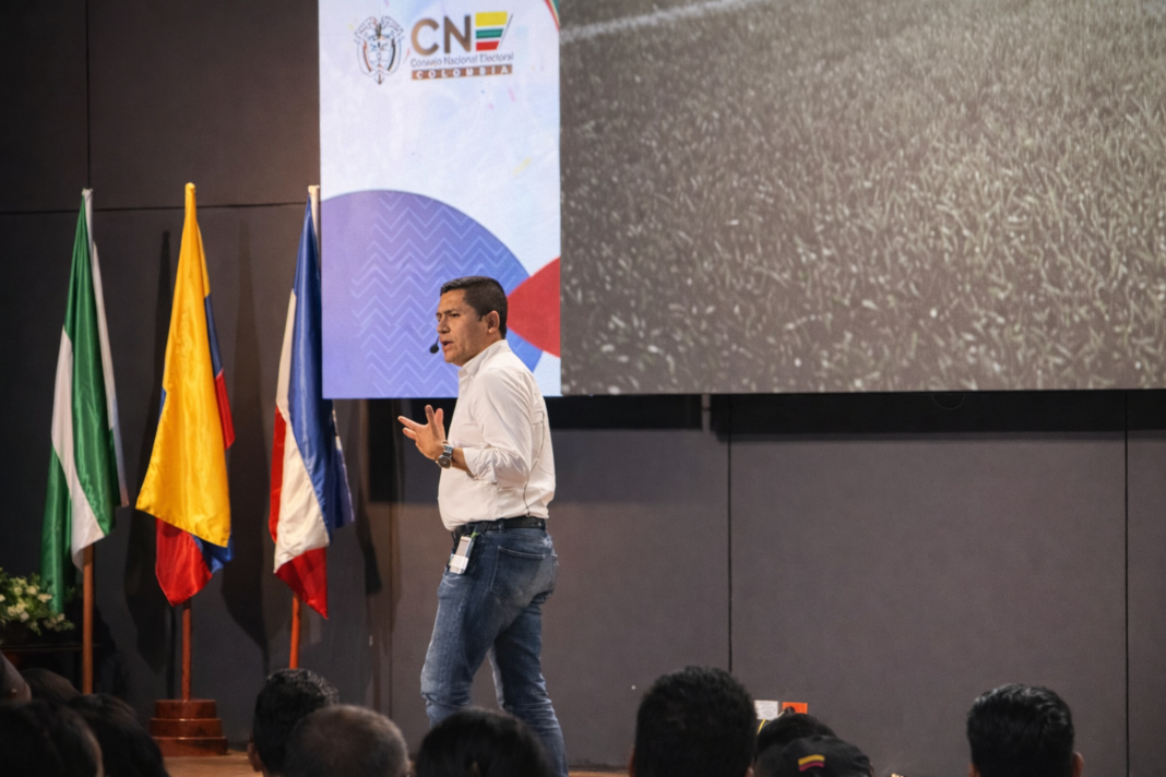 Hombre con camisa blanca y jeans dando una conferencia en un auditorio frente a una pantalla con el logo del Consejo Nacional Electoral (CNE). A la izquierda se observan las banderas del Cesar, Valledupar y Colombia.