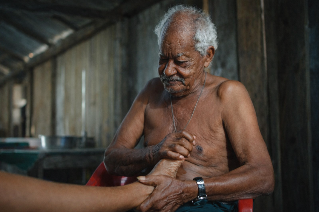 Primer plano de un sobandero tradicional, un hombre mayor, Afrodescendiente y sin camisa, concentrado mientras masajea y manipula el pie de una persona en un interior rústico de madera. Lleva un reloj negro y un collar.