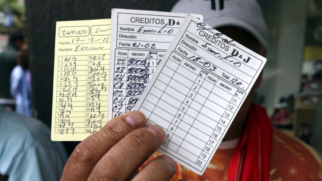 Mano sosteniendo varias tarjetas de cartulina para el control de pagos diarios de préstamos informales, con nombres, fechas y abonos escritos a mano.