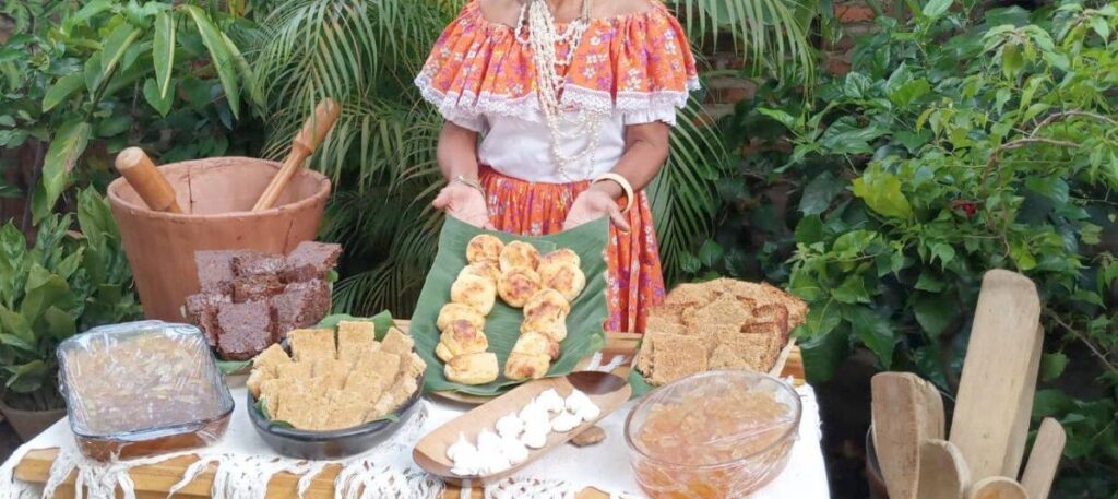 Una matrona local vestida con traje folclórico colombiano presenta una mesa repleta de diversos dulces artesanales tradicionales del Caribe y Valledupar, sobre hojas de plátano y en vasijas de barro, con un pilón al fondo.