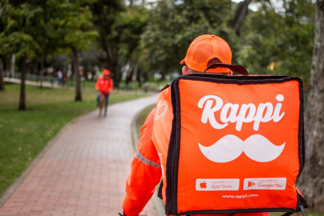 Un repartidor de Rappi, visto desde atrás, camina por un sendero pavimentado en un parque llevando una bolsa aislada de color naranja. La bolsa cuenta con el logotipo de bigote y insignias de descarga de la App Store y Google Play.