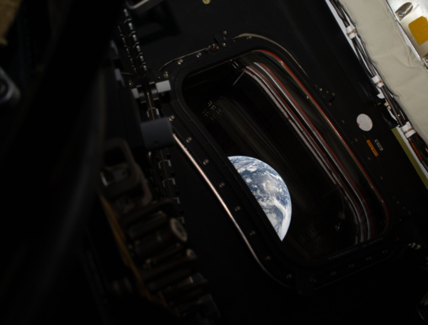 Vista íntima a través de una ventana redonda de la cápsula Orion de la misión Artemis II de la NASA, mostrando un gajo de la Tierra azul y blanca contra el negro del espacio profundo.