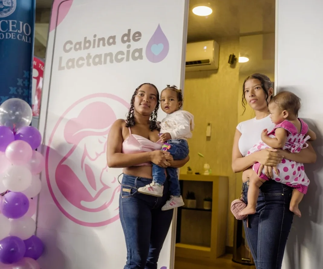 Dos madres jóvenes cargando a sus bebés frente a la entrada de la nueva Cabina de Lactancia en Cali, con decoración de globos y el logo del Concejo de Cali al fondo.