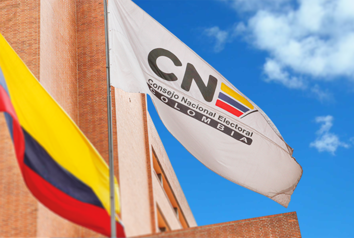 Bandera blanca del Consejo Nacional Electoral (CNE) de Colombia junto a la bandera nacional frente a un edificio de ladrillo bajo un cielo azul despejado.