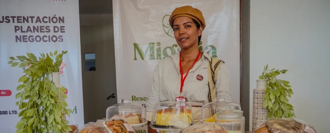 Emprendedora en Valledupar con boina marrón sonriendo frente a su stand de repostería artesanal, durante una jornada de sustentación de planes de negocios.