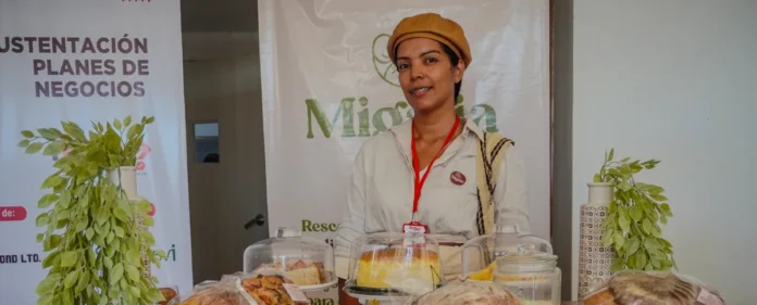 Emprendedora en Valledupar con boina marrón sonriendo frente a su stand de repostería artesanal, durante una jornada de sustentación de planes de negocios.