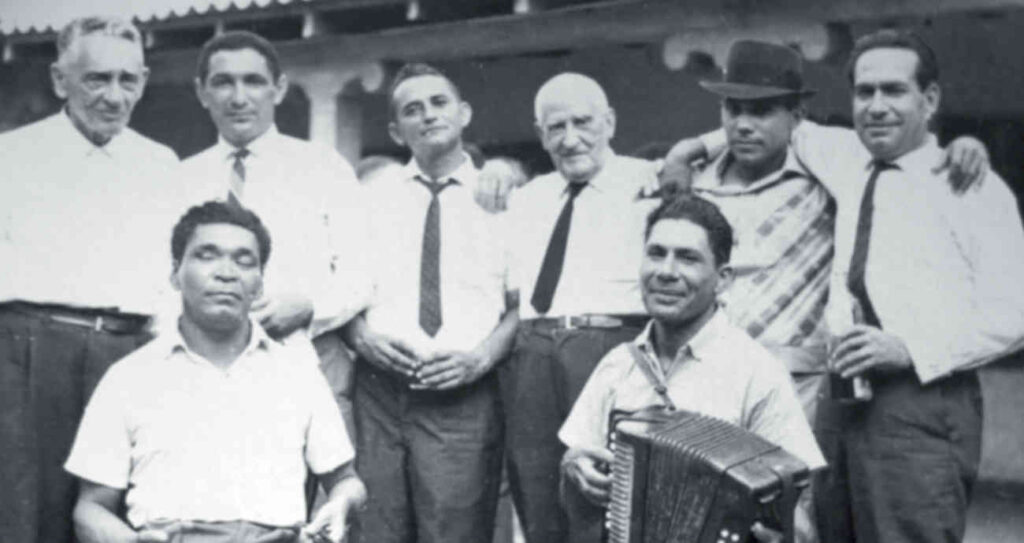 Fotografía histórica en blanco y negro de un grupo de ocho hombres de mediados del siglo XX posando juntos; un hombre en primer plano toca un acordeón mientras sonríe.