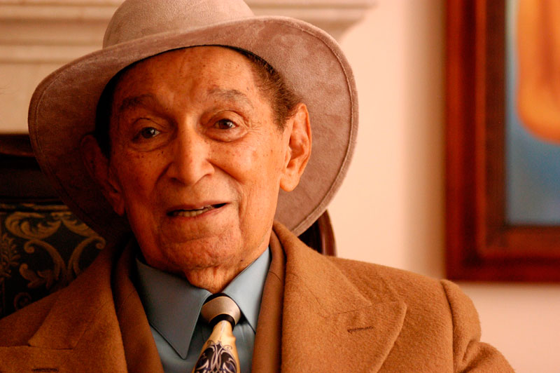 Retrato fotográfico de Rafael Escalona, el legendario compositor colombiano de vallenato, sonriendo ampliamente en un patio tradicional de Valledupar al atardecer, con un cuaderno de notas y una pluma sobre una mesa rústica y un acordeón desenfocado al fondo.