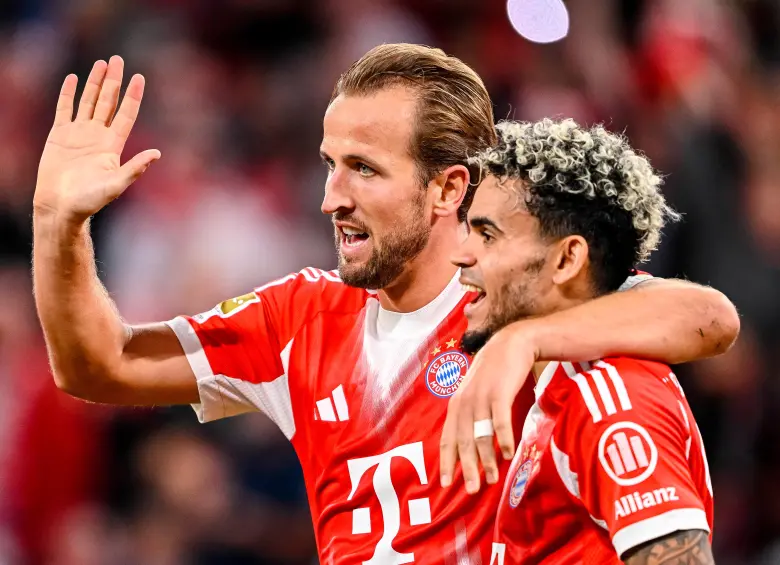 Harry Kane y Luis Díaz celebrando abrazados un gol con el uniforme rojo del Bayern Múnich. Kane levanta la mano saludando a la grada y Díaz sonríe con su característico cabello rubio crespo.