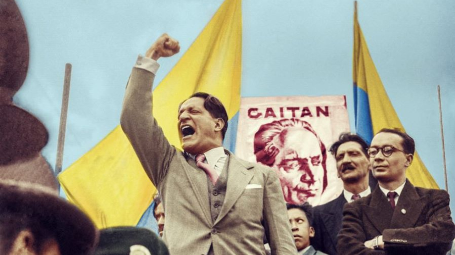 Jorge Eliécer Gaitán con el puño en alto dando un discurso frente a una multitud, con banderas amarillas y un cartel con su rostro de fondo.