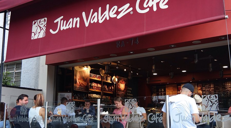 Un plano de vista lateral y detallado de la fachada de un Juan Valdez Café en la dirección 8A-74 en Bogotá, Colombia, durante el día, con clientes disfrutando de café en el exterior. La distintiva marquesina roja con el logotipo y el texto 