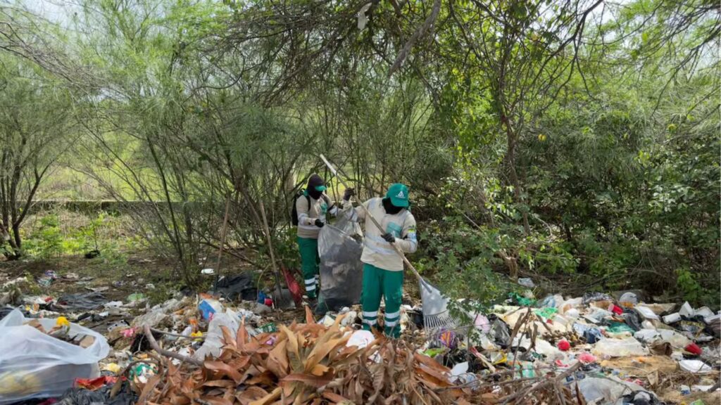 Dos operarios con uniforme verde de la Alcaldía de Riohacha recolectan basura acumulada con rastrillos y bolsas en un punto crítico ambiental de la vía Riohacha–Maicao.