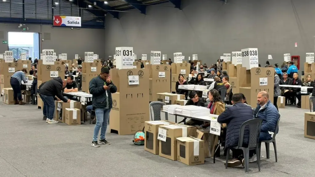 Elecciones presidenciales 41 millones habilitados: Vista general de un gran puesto de votación bajo techo en Colombia, mostrando cubículos de cartón, mesas con jurados y ciudadanos organizando la jornada electoral.