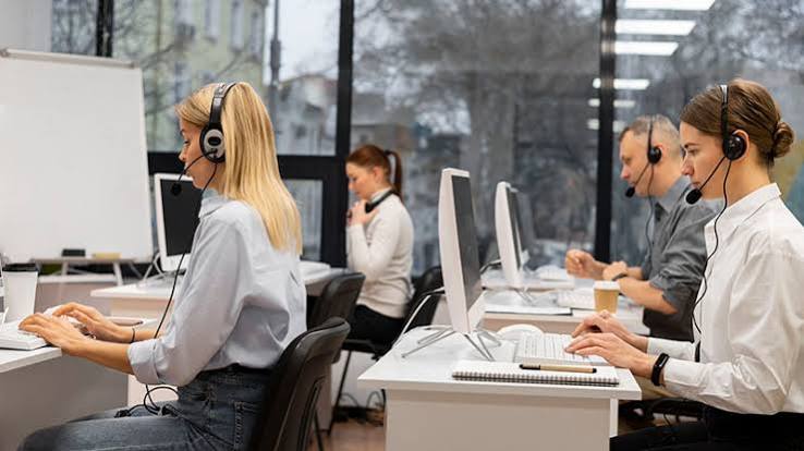 Empleados atienden sus labores de manera presencial en una oficina con equipos de cómputo y diademas.