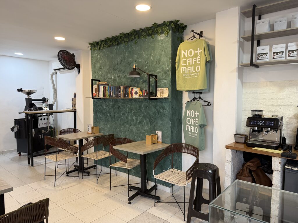 Interior de la tienda Campo Gourmet en Valledupar, con mesas para clientes, una pared decorada con libros y camisetas con el lema "No más café malo".