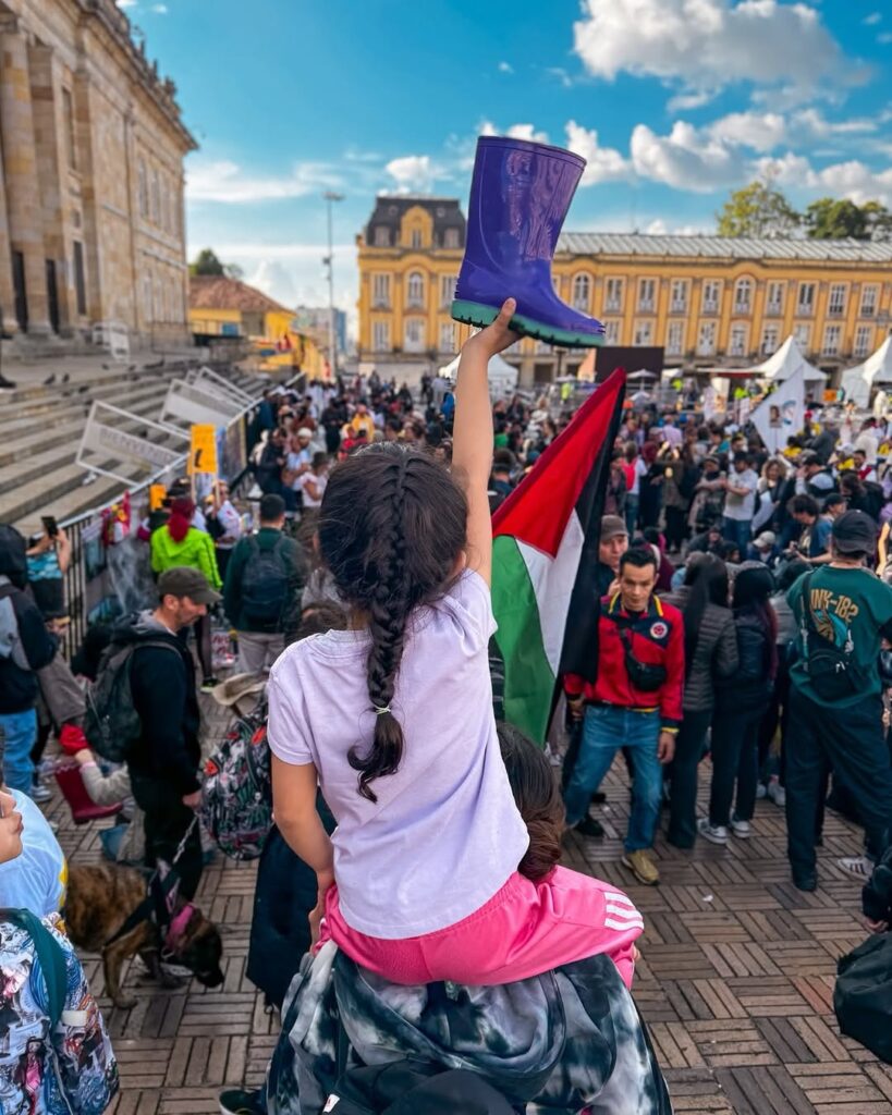 Niña sostiene una bota morada durante una manifestación en la Plaza de Bolívar de Bogotá, con una bandera palestina y decenas de asistentes al fondo.
