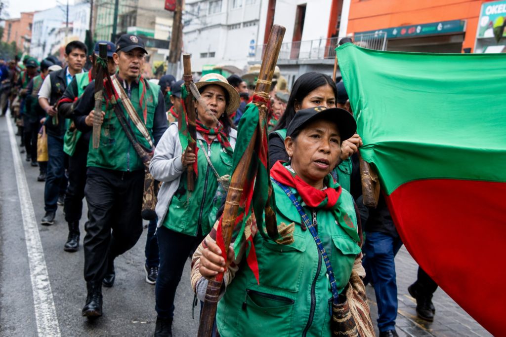 Integrantes de la Minga Indígena marchan por una vía urbana con bastones de mando y una bandera verde y roja.
