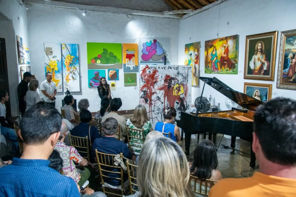 Público sentado en sillas doradas escuchando a una mujer hablar por micrófono en una galería de arte con coloridas pinturas y un piano de cola negro durante ArtVa 2025.