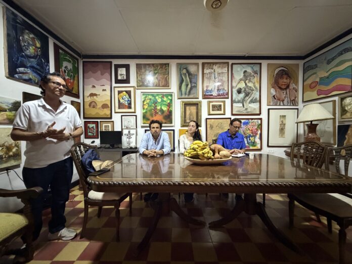 Un hombre de pie y tres personas sentadas en una mesa de madera, reunidos en una sala con paredes cubiertas de múltiples cuadros y pinturas, preparándose para la Feria de Arte ARTVA Inmersión 2026