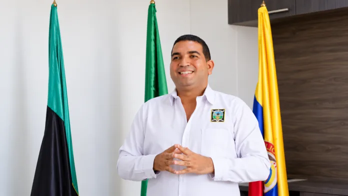José José Romero Carrillo con camisa blanca y tres banderas al fondo en una oficina.
